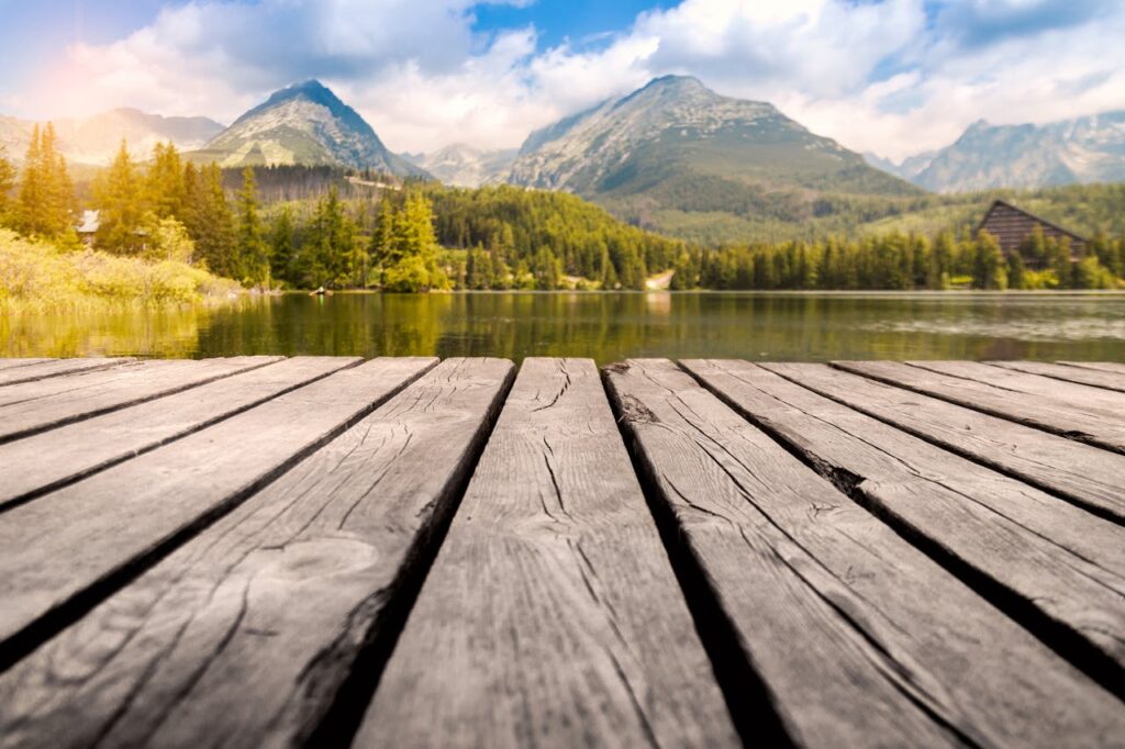 Calm mountain lake with a wooden dock, surrounded by lush forests and mountains under a clear sky.